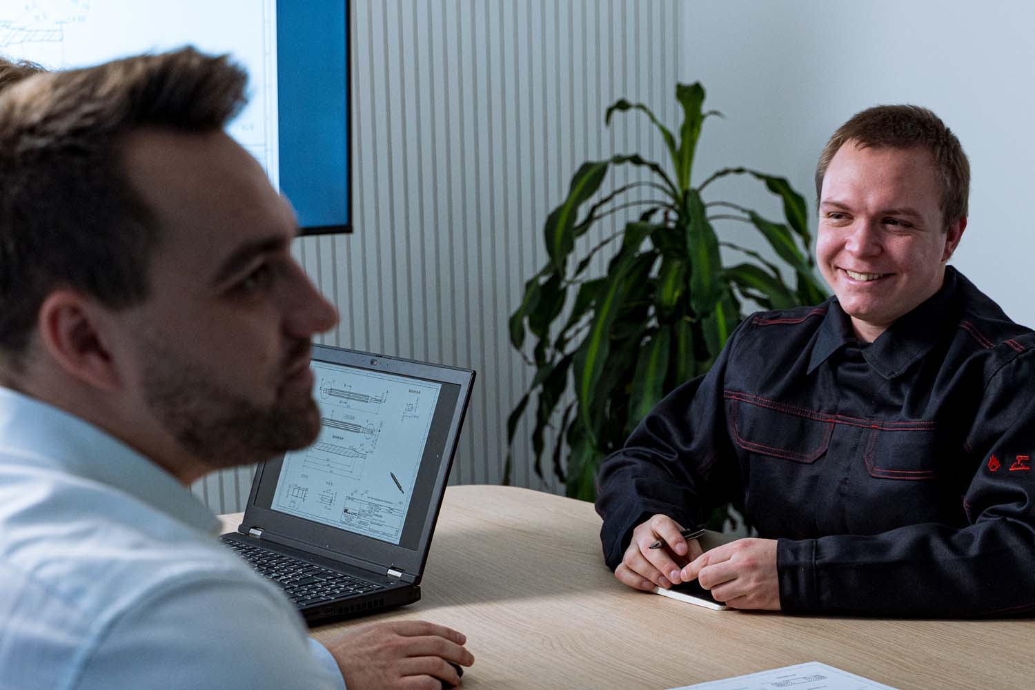 Drei Personen an einem Schreibtisch. Eine Person ist verdeckt, man erkennt, dass sie im Hintergrund eine Konstrukltionszeichung auf dem Laptop geöffnet hat. Ein Mitarbeiter sitzt gegenüber mit Schreibblock und grinst.